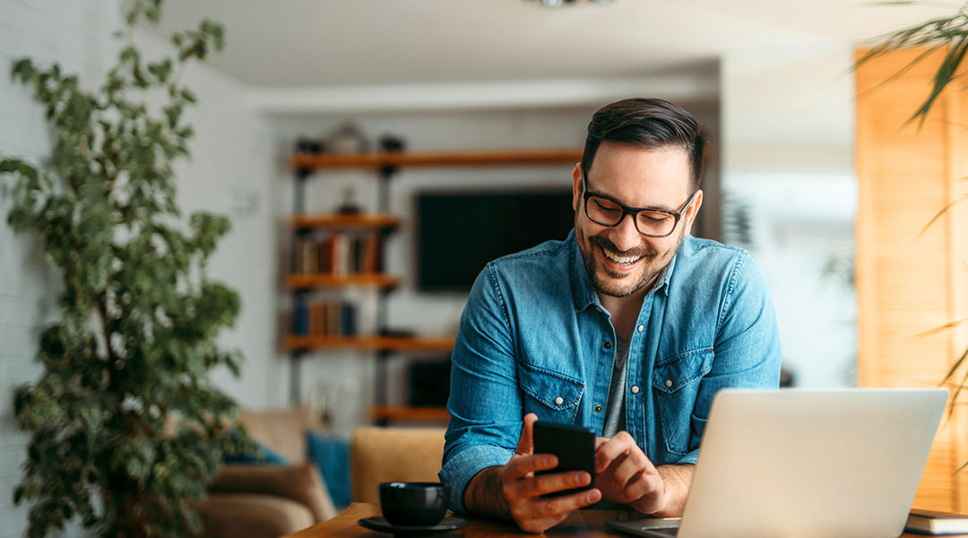 Man with glasses smiling and looking down at his phone with his laptop to his left